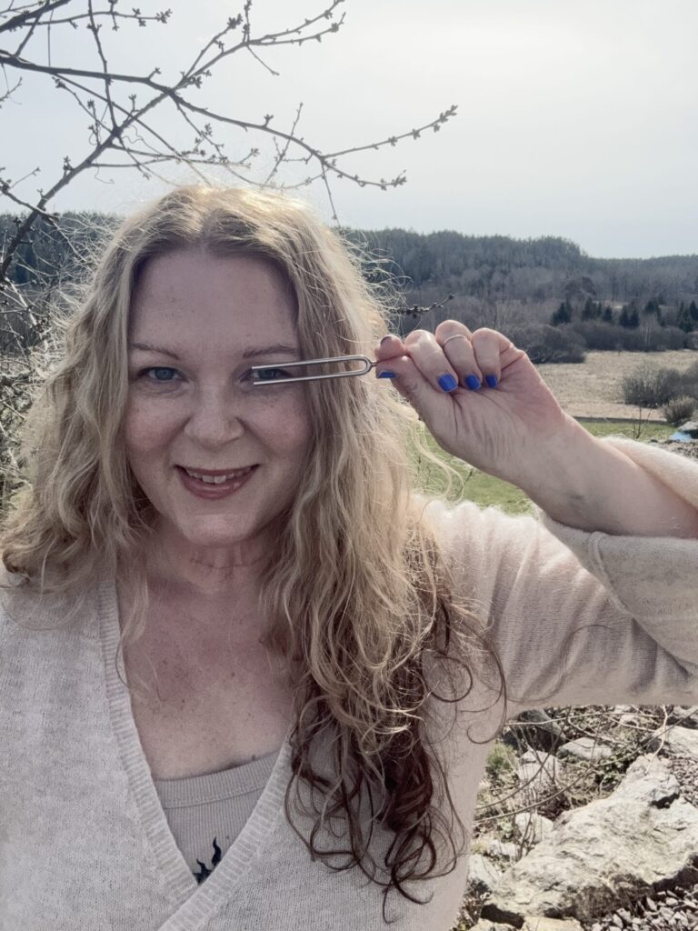 Leende porträtt av sångerskan Karin Ericsson Back som håller en stämgaffel framför sig utomhus med natur i bakgrunden.