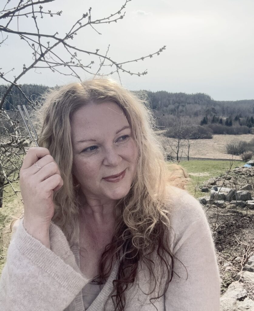 Sångerskan Karin Ericsson Back som håller i en stämgaffel utomhus med natur i bakgrunden.